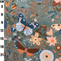 A close-up of a woven fabric with a detailed botanical and insect pattern, featuring butterflies, moths, flowers, and leaves in muted blue, orange, pink, and brown tones on a dusty teal-gray background. A black measurement scale in centimeters is printed along the left edge.