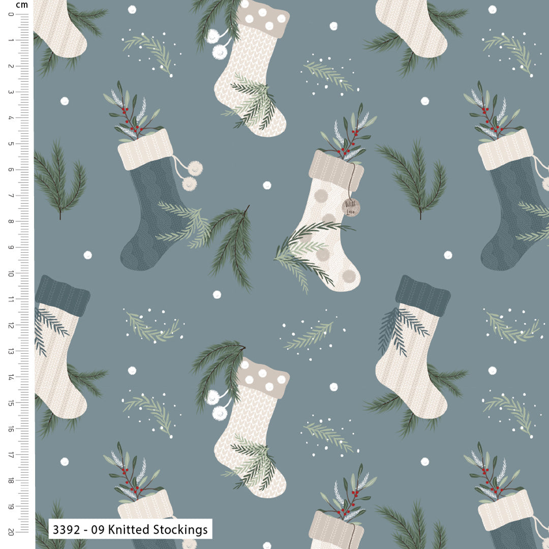 A repeating Christmas pattern on a muted blue-gray background featuring knitted stockings in cream, beige, and dark blue-gray, with pine branches, white berries, and small white dots. A white ruler along the left edge shows the scale in centimeters, and a label at the bottom left reads '3392 - 09 Knitted Stockings'.