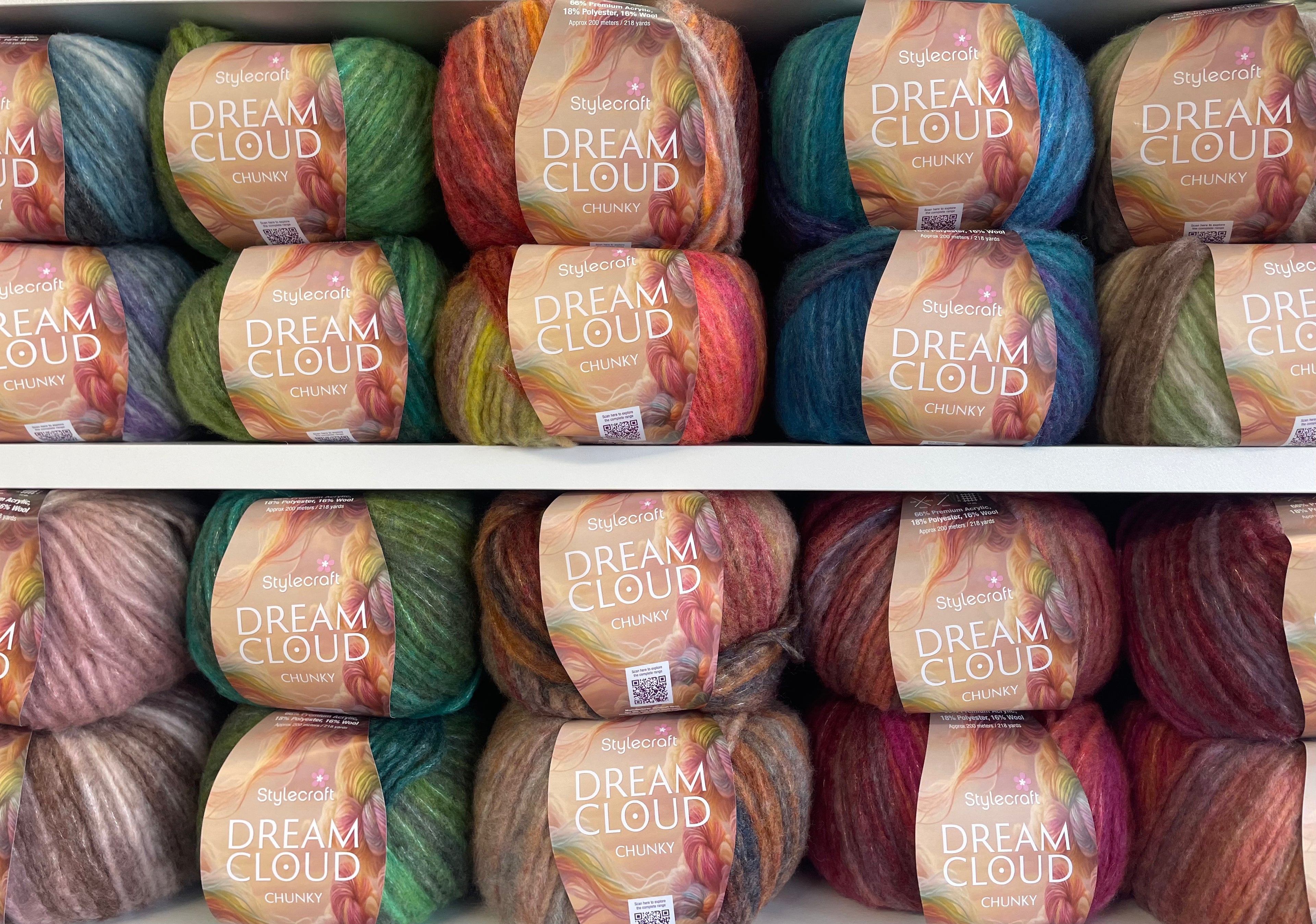 Multiple skeins of yarn arranged on white shelves, each wrapped with a label reading 'Dream Cloud Creamy.' The yarns show varied colorways including blue, green, orange, pink, purple, and brown, making the overall color identity multicolor.