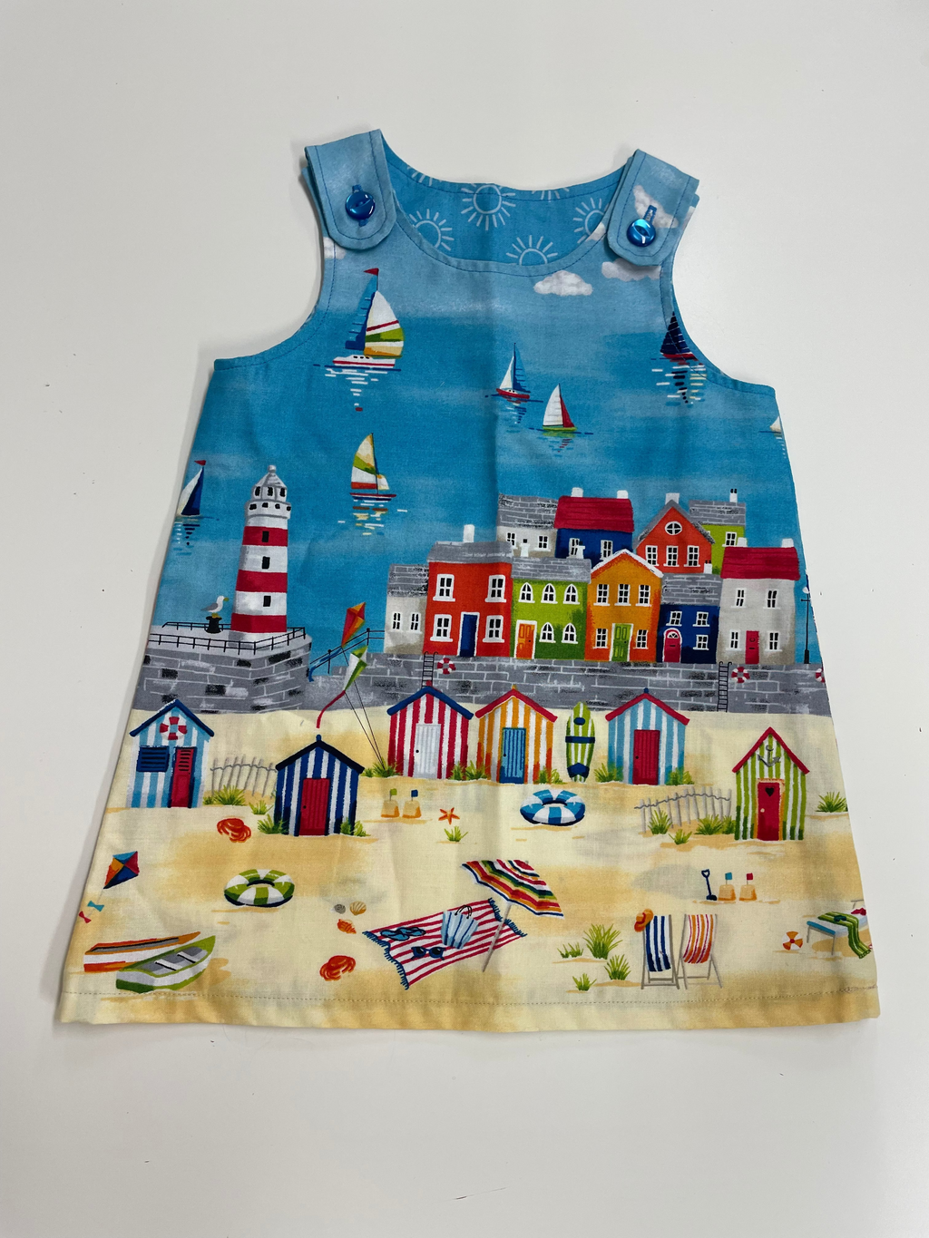 A sleeveless baby dress featuring a colorful coastal beach town print with sailboats, a lighthouse, houses, and striped beach huts on a light gray background.