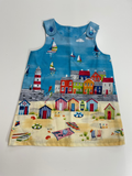 A sleeveless baby dress featuring a colorful coastal beach town print with sailboats, a lighthouse, houses, and striped beach huts on a light gray background.