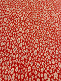 A close-up of a red-orange fabric with a repeating small white and light purple ikat-style floral pattern.