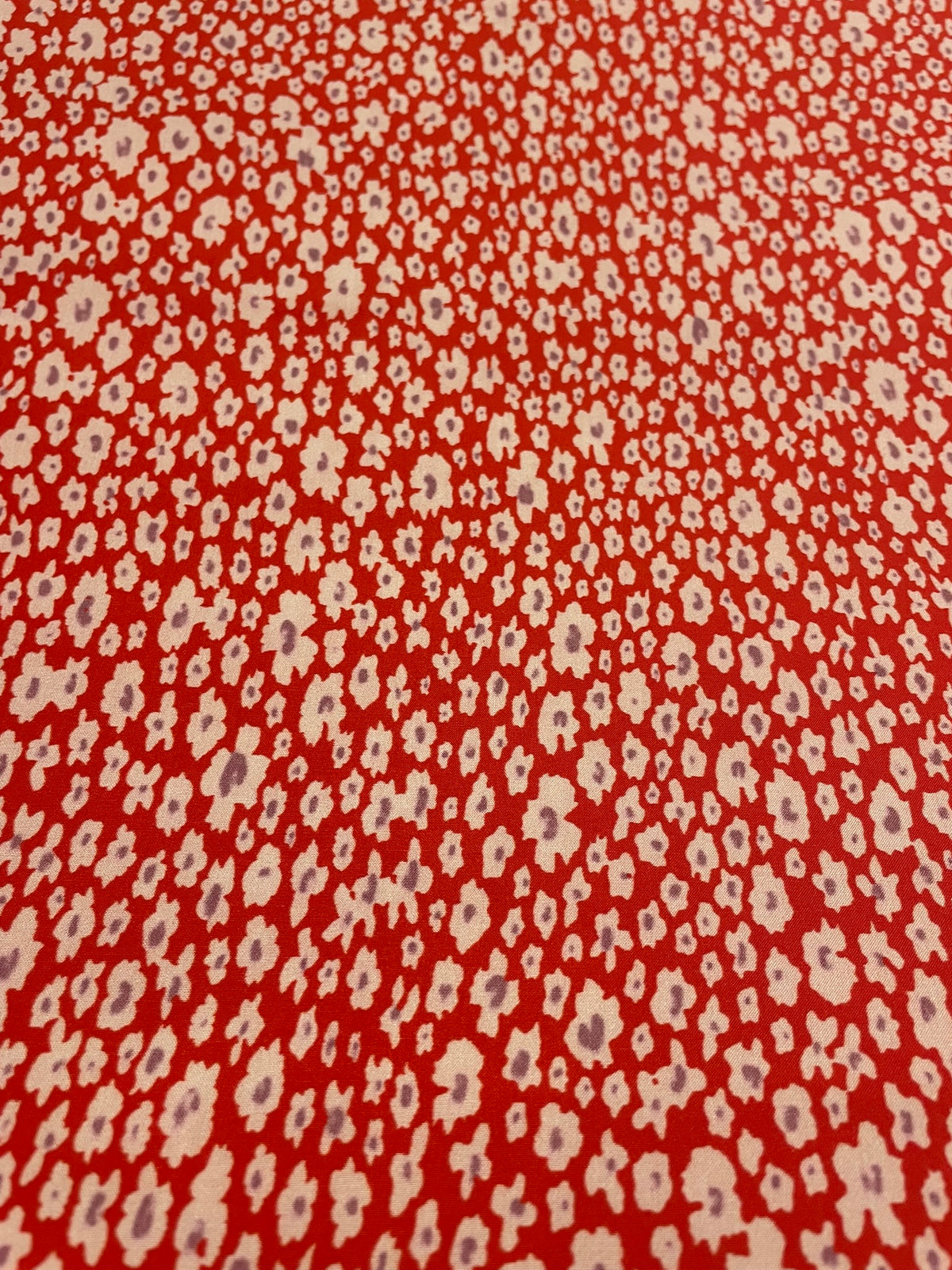 A close-up of a red-orange fabric with a repeating small white and light purple ikat-style floral pattern.