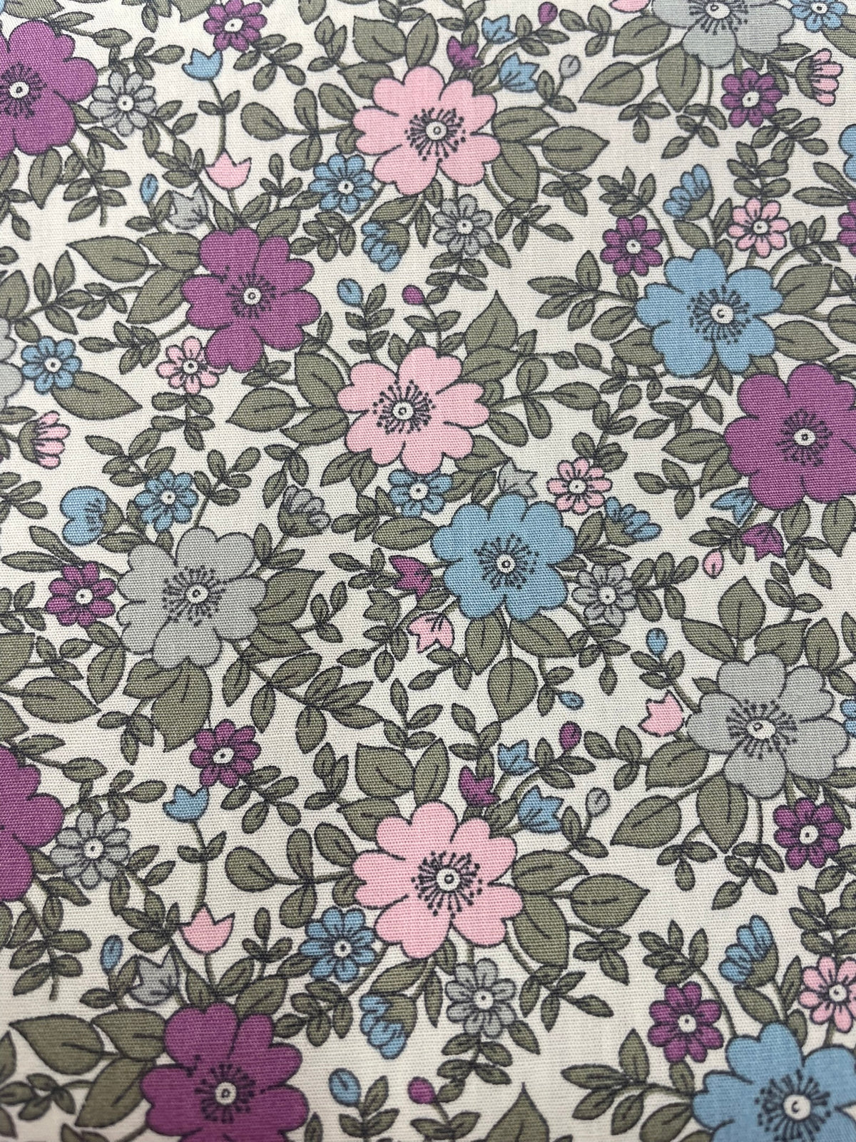 A close-up of a floral fabric pattern featuring pink, purple, and blue flowers with gray-green leaves on a cream background.