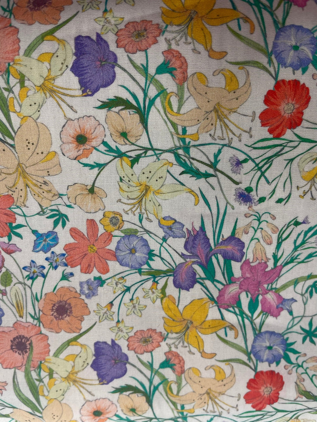 A close-up of a fabric pattern featuring a dense, multicolor floral print with lilies, poppies, and wildflowers on a cream-colored background.