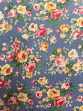 A close-up of a floral fabric pattern featuring pink, yellow, and white roses with green leaves on a dusty blue background.