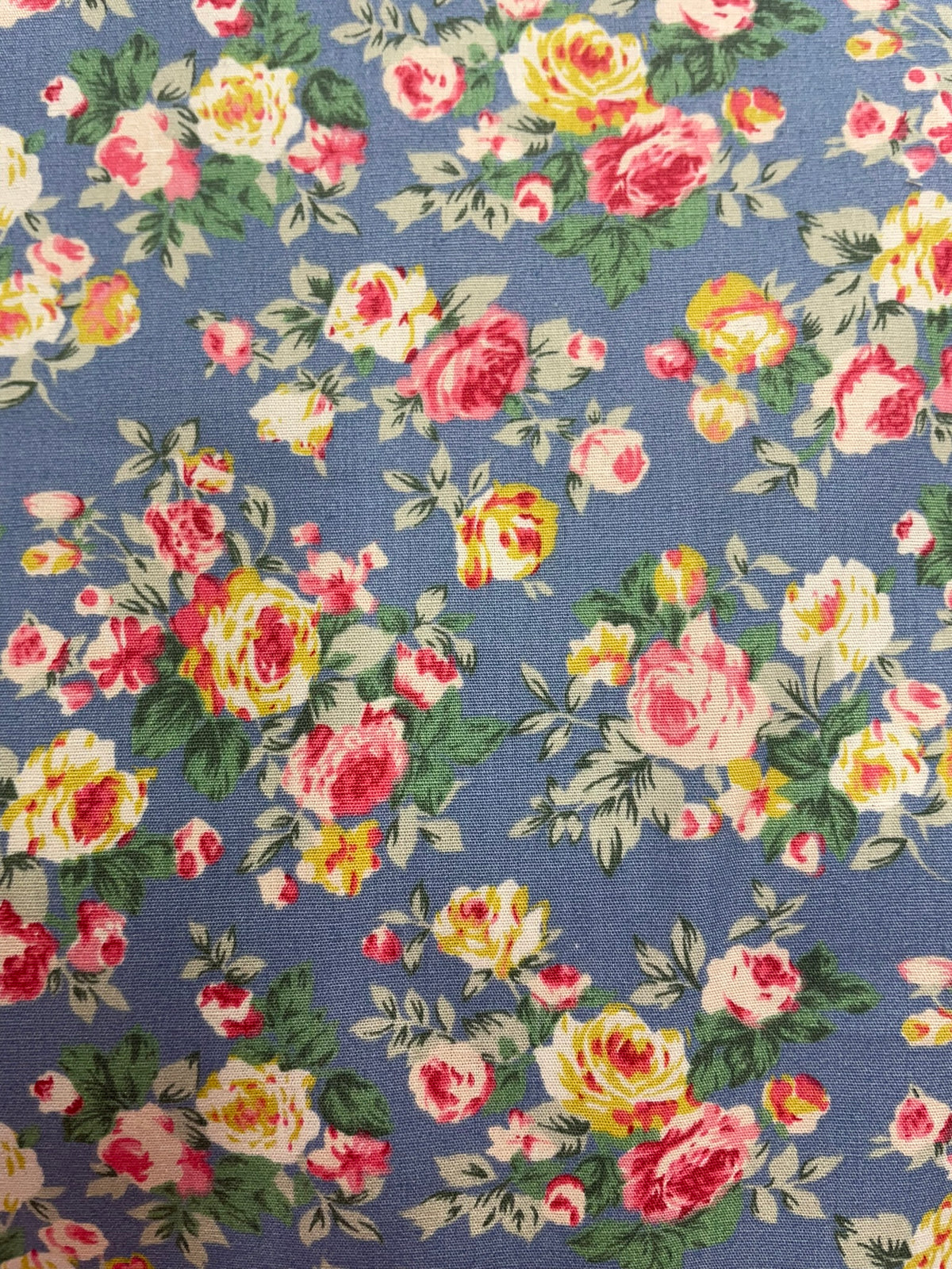 A close-up of a floral fabric pattern featuring pink, yellow, and white roses with green leaves on a dusty blue background.
