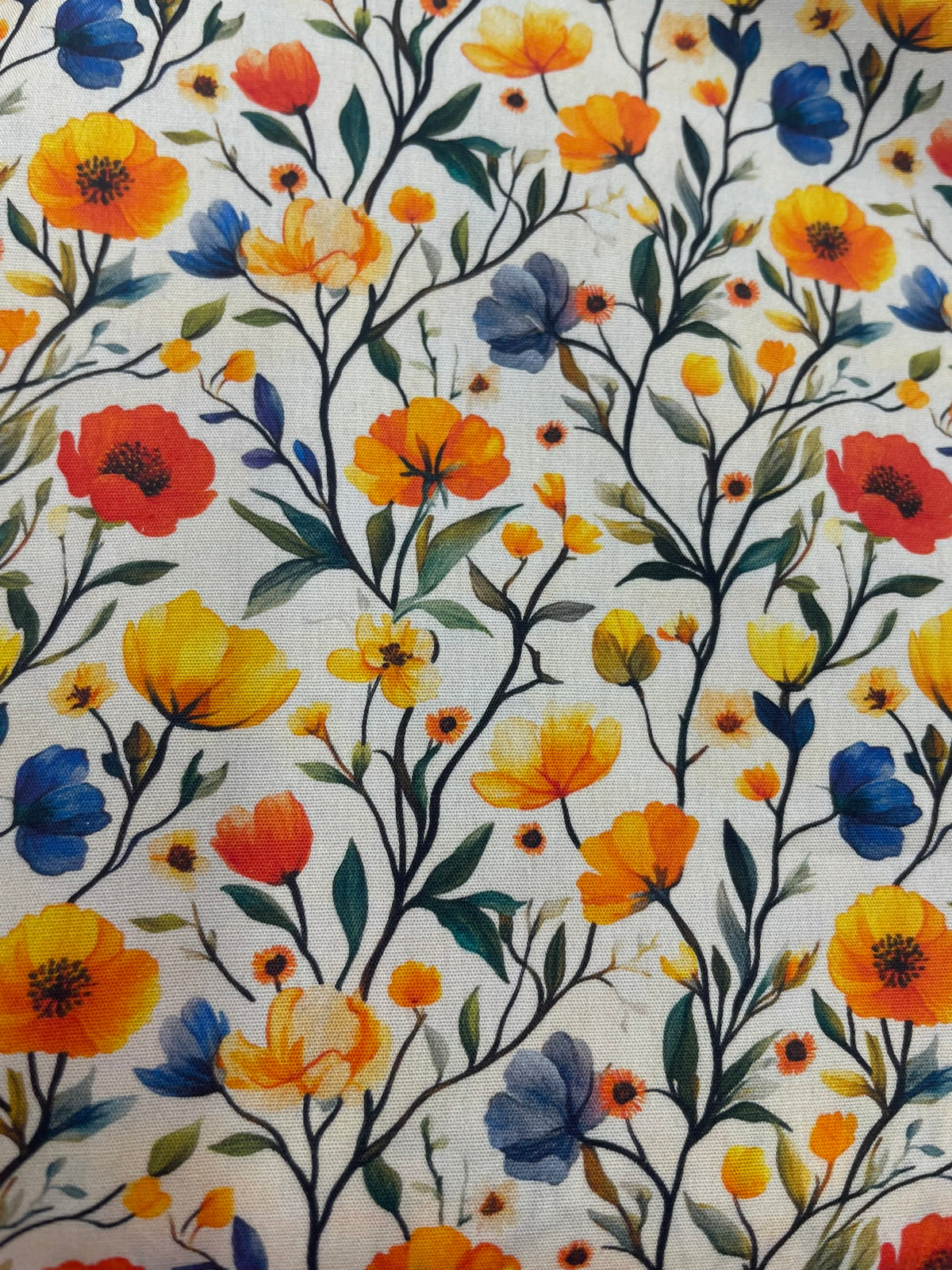 A close-up of a fabric pattern featuring a dense, colorful wildflower print on a cream background. The design includes orange, yellow, red, and blue flowers with green and dark blue leaves and stems.