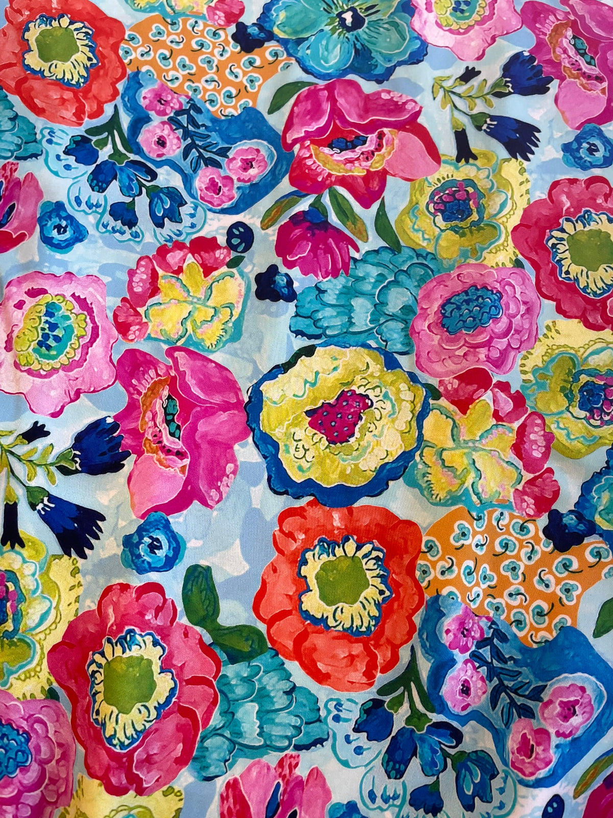 A close-up of a colorful floral patterned fabric featuring large, vibrant hand-painted style flowers in pink, red, yellow, blue, and green on a light blue background.
