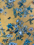 A close-up of a mustard yellow fabric with an all-over floral print in shades of blue, white, and dark navy, featuring botanical motifs and small butterflies.