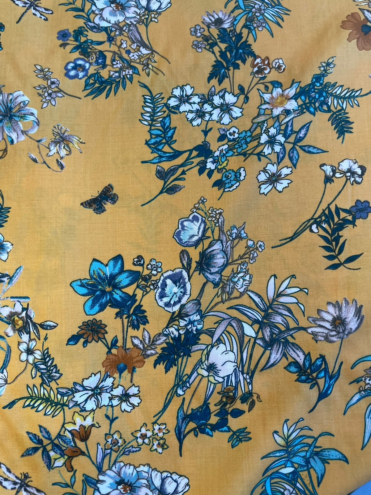 A close-up of a mustard yellow fabric with an all-over floral print in shades of blue, white, and dark navy, featuring botanical motifs and small butterflies.