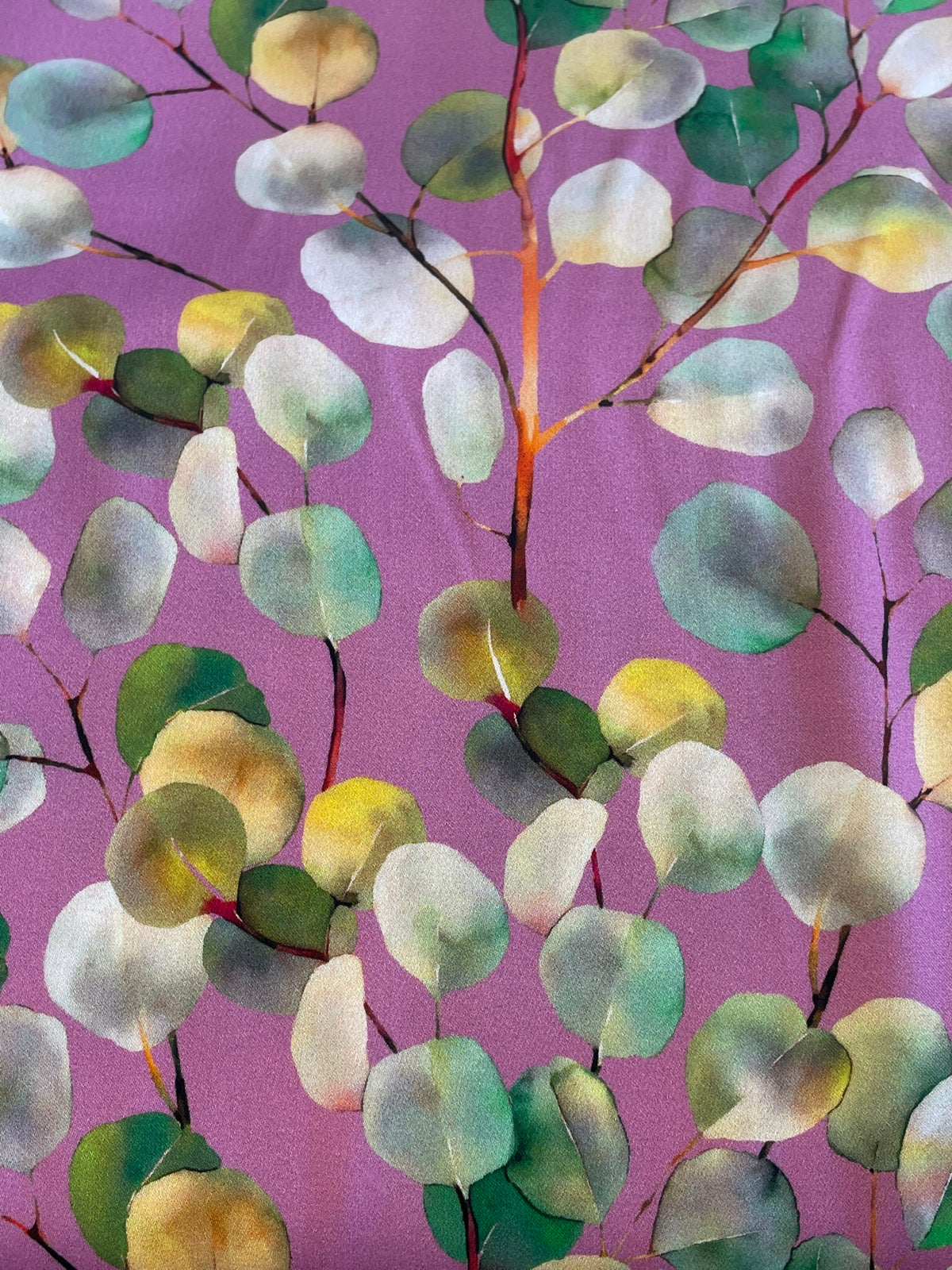 A close-up of a fabric pattern featuring watercolor-style eucalyptus leaves in shades of green, yellow, and white on a dusty purple background.