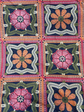 A close-up of a crocheted textile featuring a repeating granny square pattern with pink and orange floral motifs on dark navy backgrounds, bordered by green and white crochet details.
