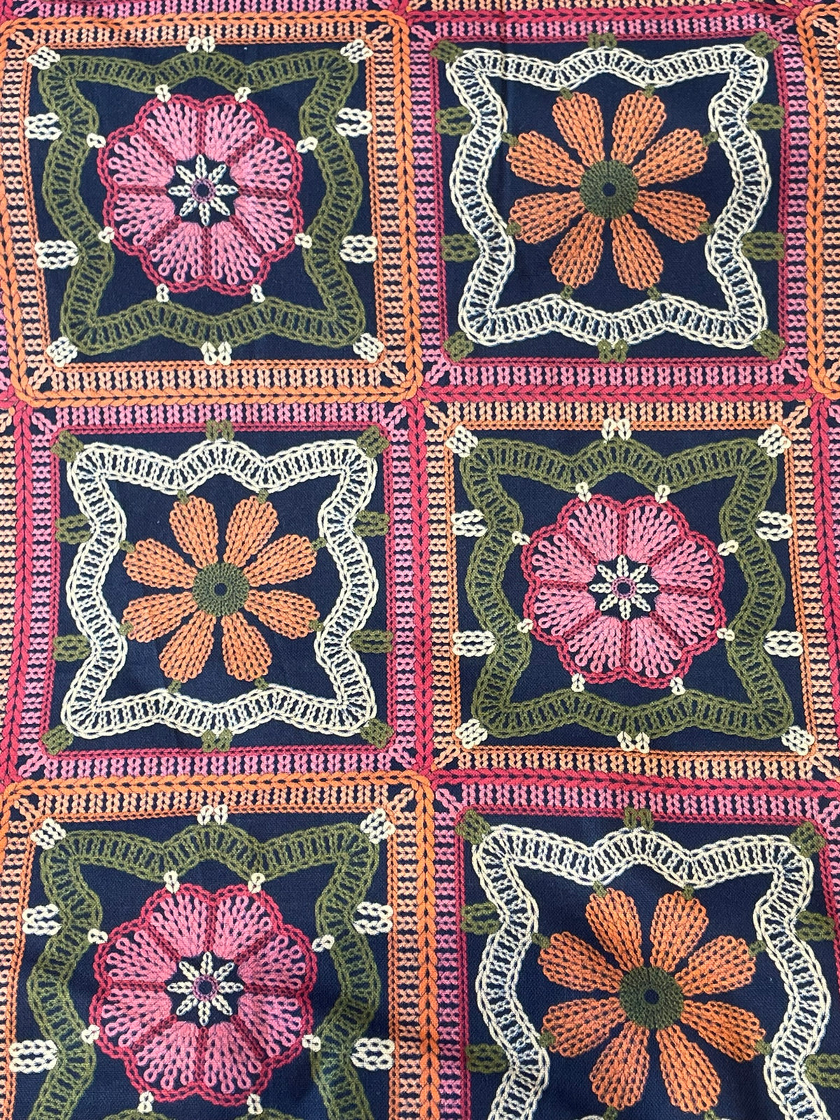 A close-up of a crocheted textile featuring a repeating granny square pattern with pink and orange floral motifs on dark navy backgrounds, bordered by green and white crochet details.