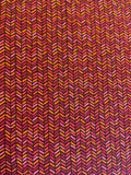 A close-up of a woven fabric with a repeating chevron or herringbone pattern in dark red, burgundy, pink, orange, and yellow.