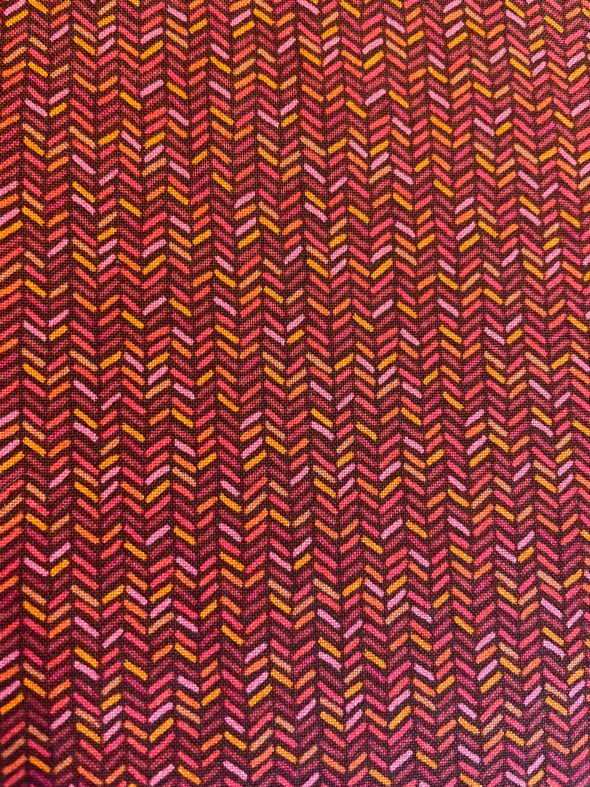 A close-up of a woven fabric with a repeating chevron or herringbone pattern in dark red, burgundy, pink, orange, and yellow.