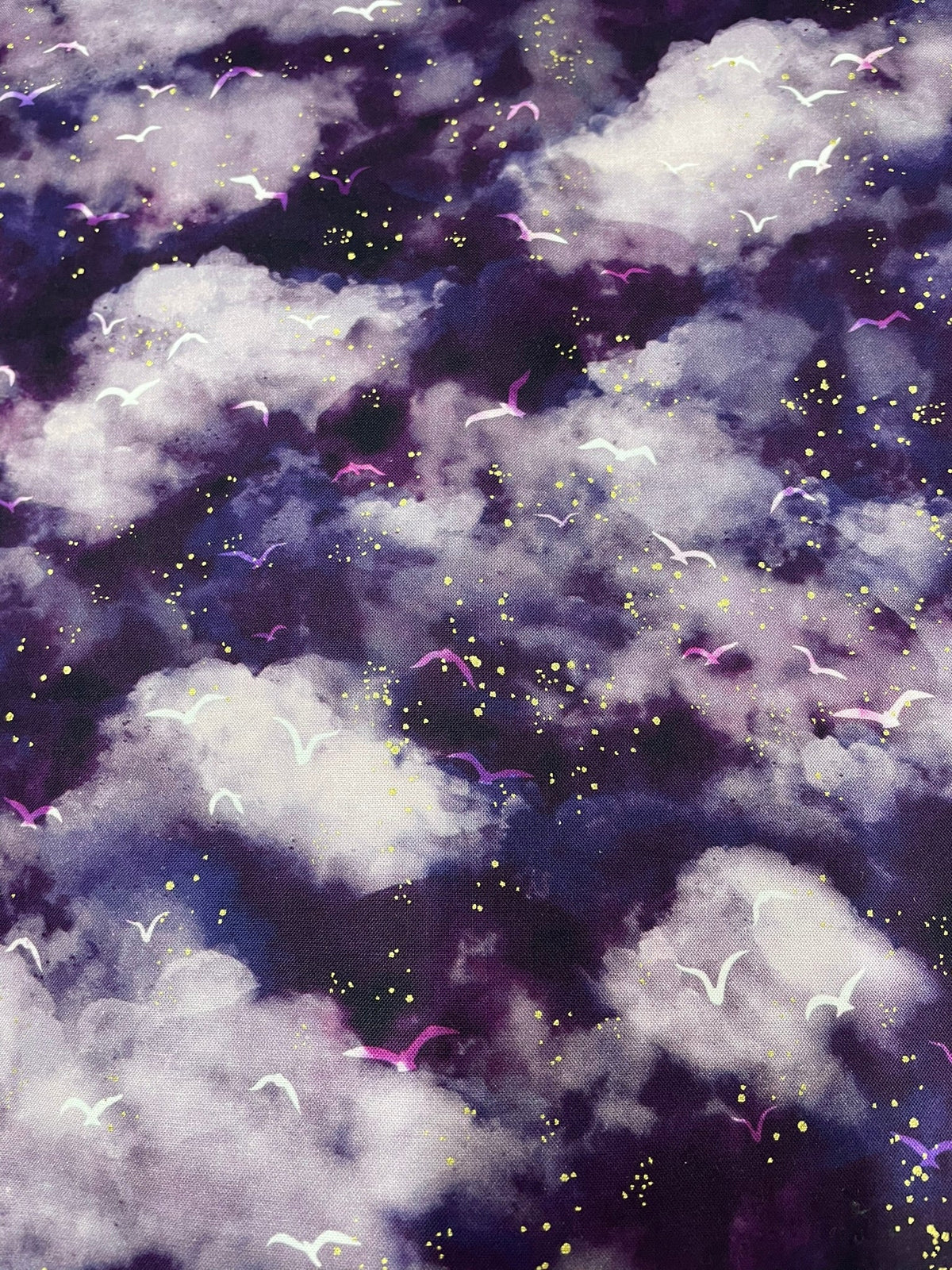 A close-up of a fabric pattern featuring a dark purple and black cloudy background with white and gray clouds, small yellow glitter-like dots, and pink and purple abstract curved accents.