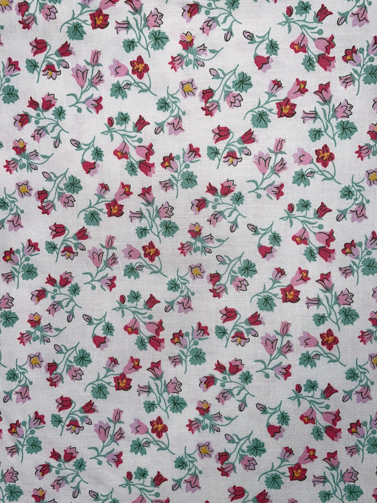 A close-up of a white fabric with a repeating floral pattern featuring pink, red, and green flowers with yellow centers.