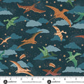 A fabric pattern featuring colorful prehistoric flying dinosaurs, including a large brown one in the center, flying over blue clouds and small gold stars on a dark teal night sky. A white ruler with the andover brand is shown along the bottom edge.