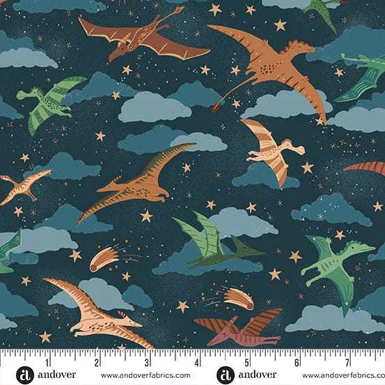 A fabric pattern featuring colorful prehistoric flying dinosaurs, including a large brown one in the center, flying over blue clouds and small gold stars on a dark teal night sky. A white ruler with the andover brand is shown along the bottom edge.