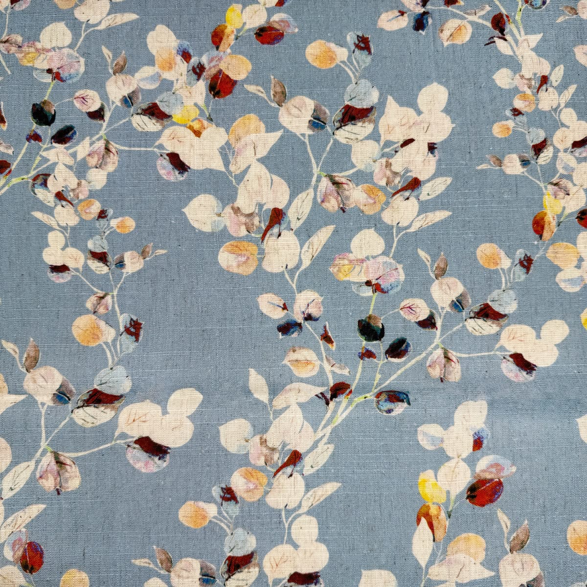A close-up of a fabric pattern featuring a delicate, painterly floral design of eucalyptus-like leaves in cream, beige, rust red, mustard yellow, and dark brown on a muted slate blue-gray background.
