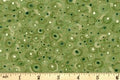 A close-up of a fabric pattern featuring concentric circles in shades of green, olive, and dark green on a light beige background, with a wooden ruler at the bottom showing the scale.