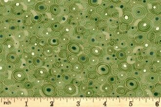 A close-up of a fabric pattern featuring concentric circles in shades of green, olive, and dark green on a light beige background, with a wooden ruler at the bottom showing the scale.